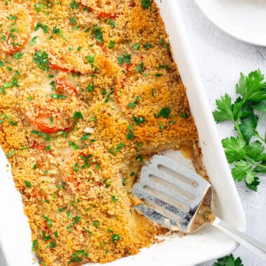 A potato-tomato casserole with Gruyere in a white casserole dish, flanked by a spatula and some sprigs of fresh parsley.