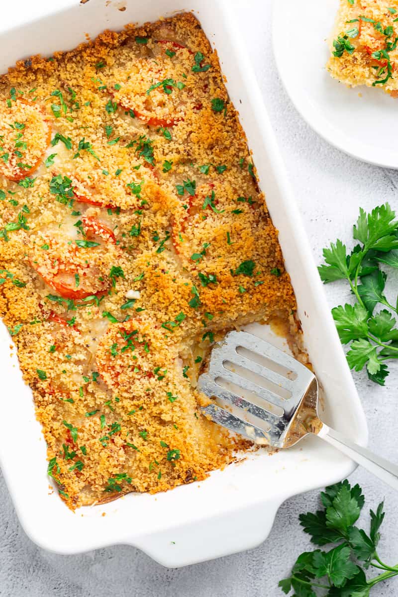 A potato-tomato casserole with Gruyere in a white casserole dish, flanked by a spatula and some sprigs of fresh parsley.