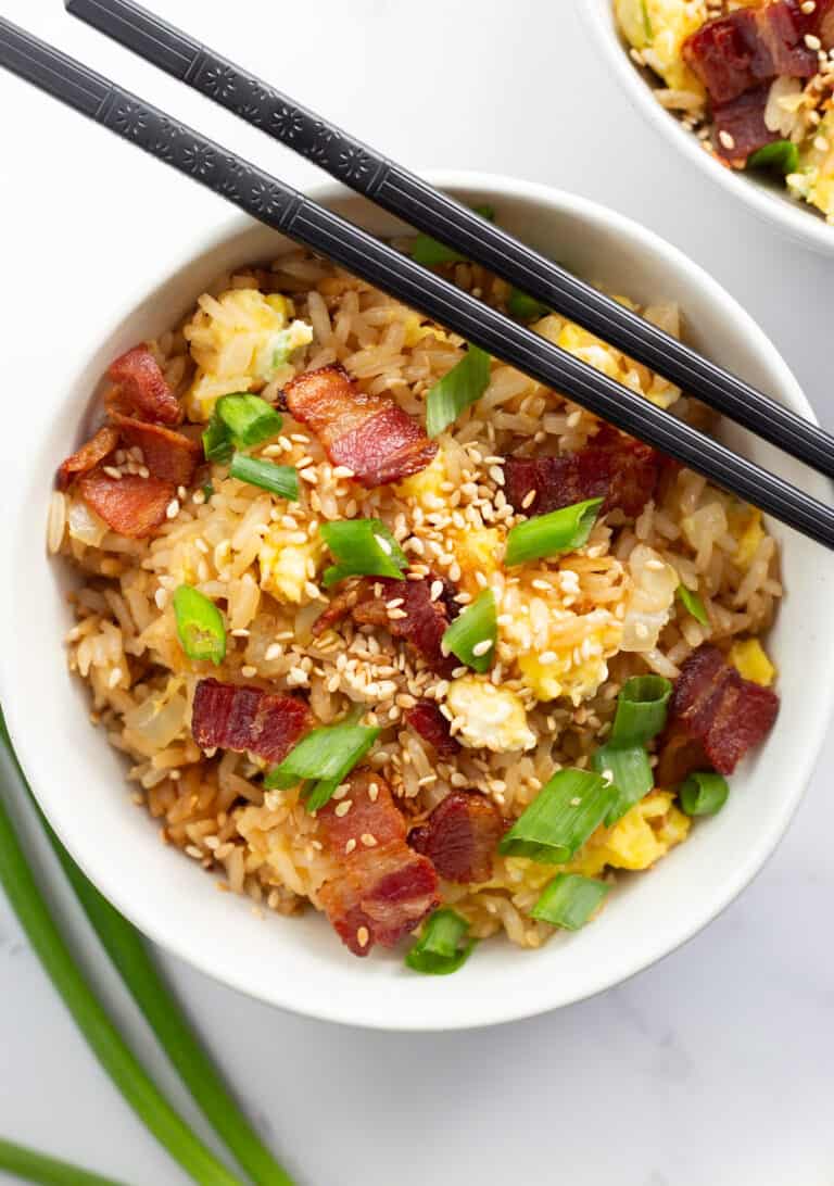 Bacon and egg fried rice in a white bowl with chopsticks resting on top of it.