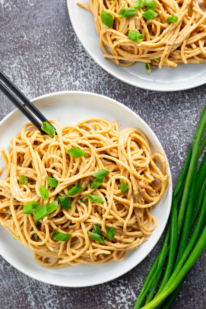 Plate of cold sesame noodles garnished with scallions, with a bushel of whole scallions laying beside it.