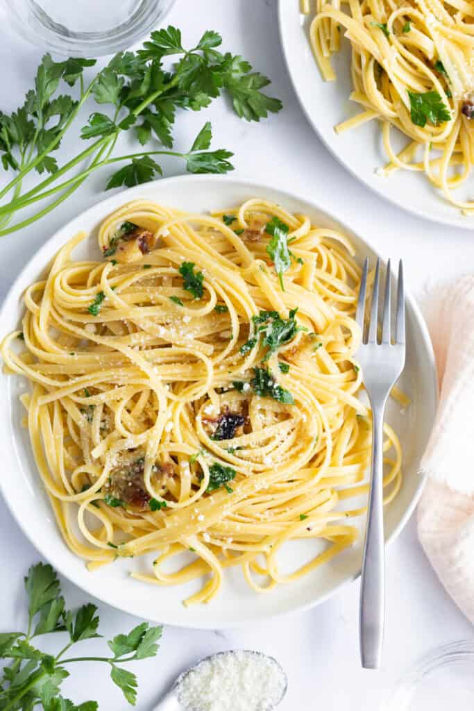 Two plates of linguine aglio e olio with roasted garlic, surrounded by sprigs of fresh parsley.