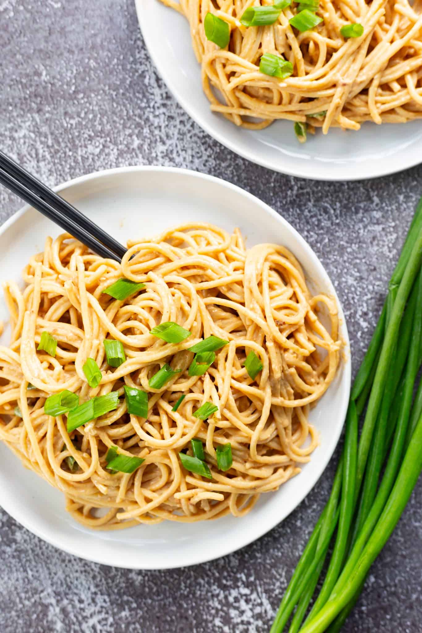 Two plates of cold sesame noodles garnished with sliced scallions, with a bushel of scallions next to one plate.