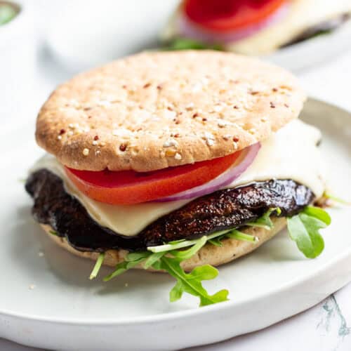 A grilled portobello burger topped with sliced tomato, sliced red onion, and melted baby Swiss on a bed of arugula and between two sandwich buns.