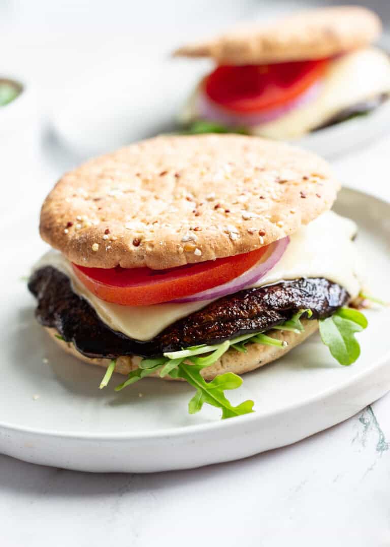 A grilled portobello burger topped with sliced tomato, sliced red onion, and melted baby Swiss on a bed of arugula and between two sandwich buns.