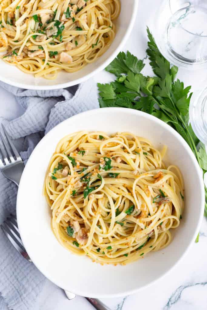 Two bowls of linguine with clams, anchovies, and breadcrumbs, with a bushel of fresh parsley placed next to one of the bowls.