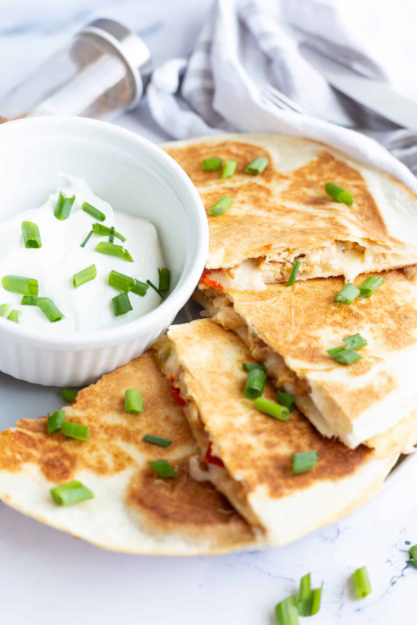 A plate of chicken quesadillas cut into four hand-held pieces and sitting next to a small bowl of sour cream garnished with scallions.