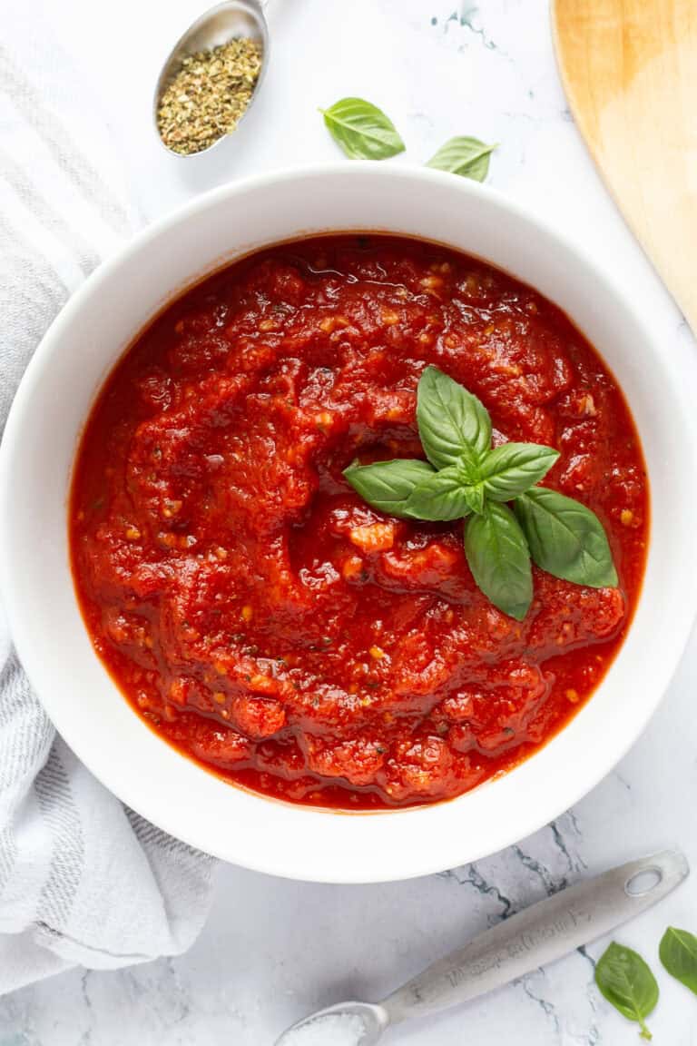 Homemade pizza sauce filling a small white bowl, with some fresh basil leaves and a wooden spoon nearby.