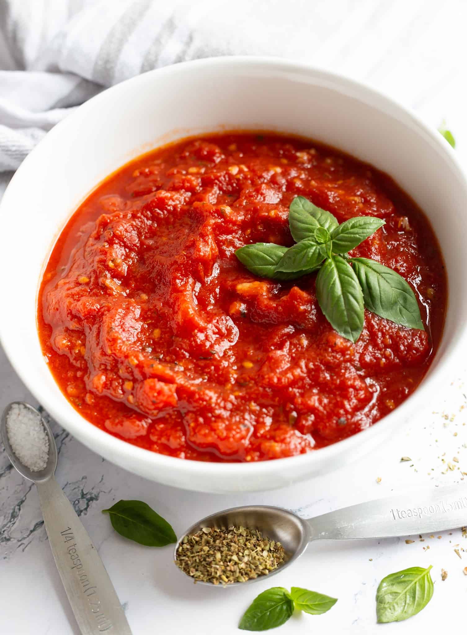 A bowl of homemade pizza sauce garnished with fresh basil, with some basil leaves and measuring spoons containing salt and dried oregano nearby.