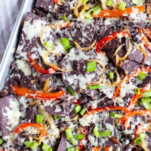 Veggie nachos sitting in a large sheet pan and covered with white cheddar cheese, black beans, cooked peppers and onions, and scallions.