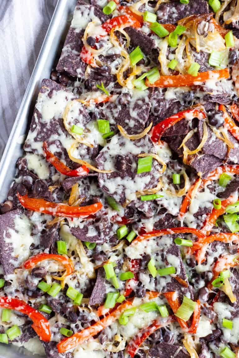 Veggie nachos sitting in a large sheet pan and covered with white cheddar cheese, black beans, cooked peppers and onions, and scallions.