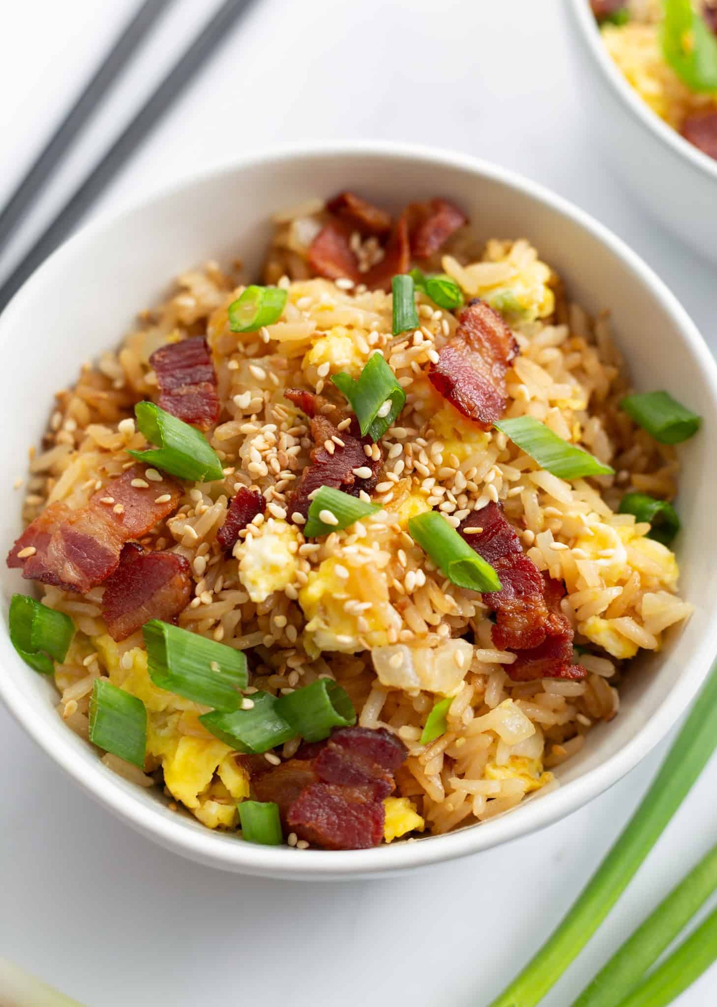 A bowl of fried rice made with bacon and eggs and topped with chopped scallions and toasted sesame seeds.