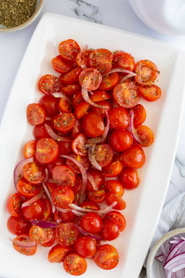 A white serving platter of marinated tomatoes with sliced red onion and dried oregano.