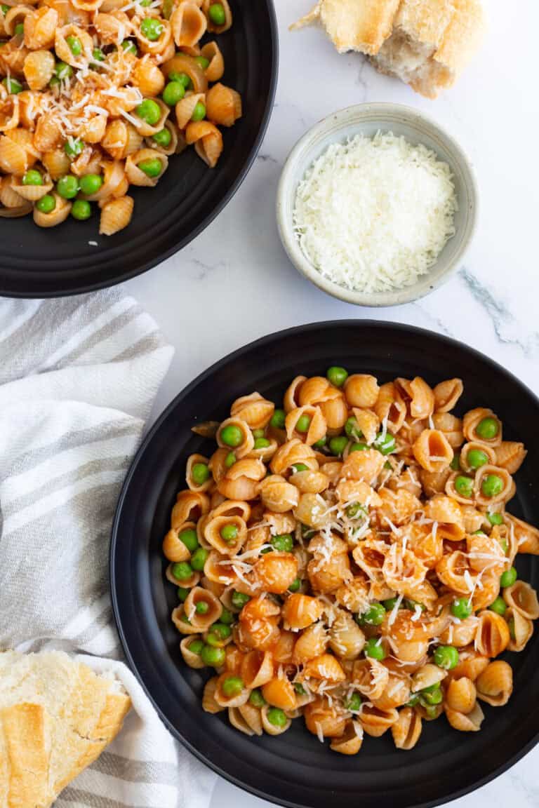 Two plates filled with pasta and peas in red sauce, accompanied by some torn Italian bread and a small bowl of grated Pecorino.