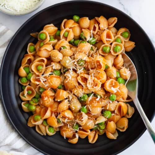 A dark plate filled with pasta and peas with red sauce, sprinkled with Parmesan. Some torn crusty bread and a small bowl of grated Parmesan are placed next to it.