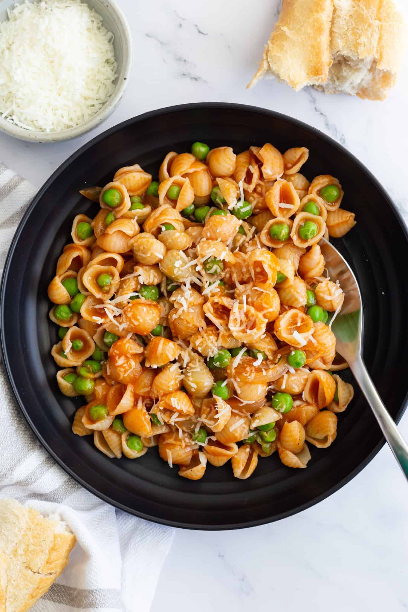 A dark plate filled with pasta and peas with red sauce, sprinkled with Parmesan. Some torn crusty bread and a small bowl of grated Parmesan are placed next to it.