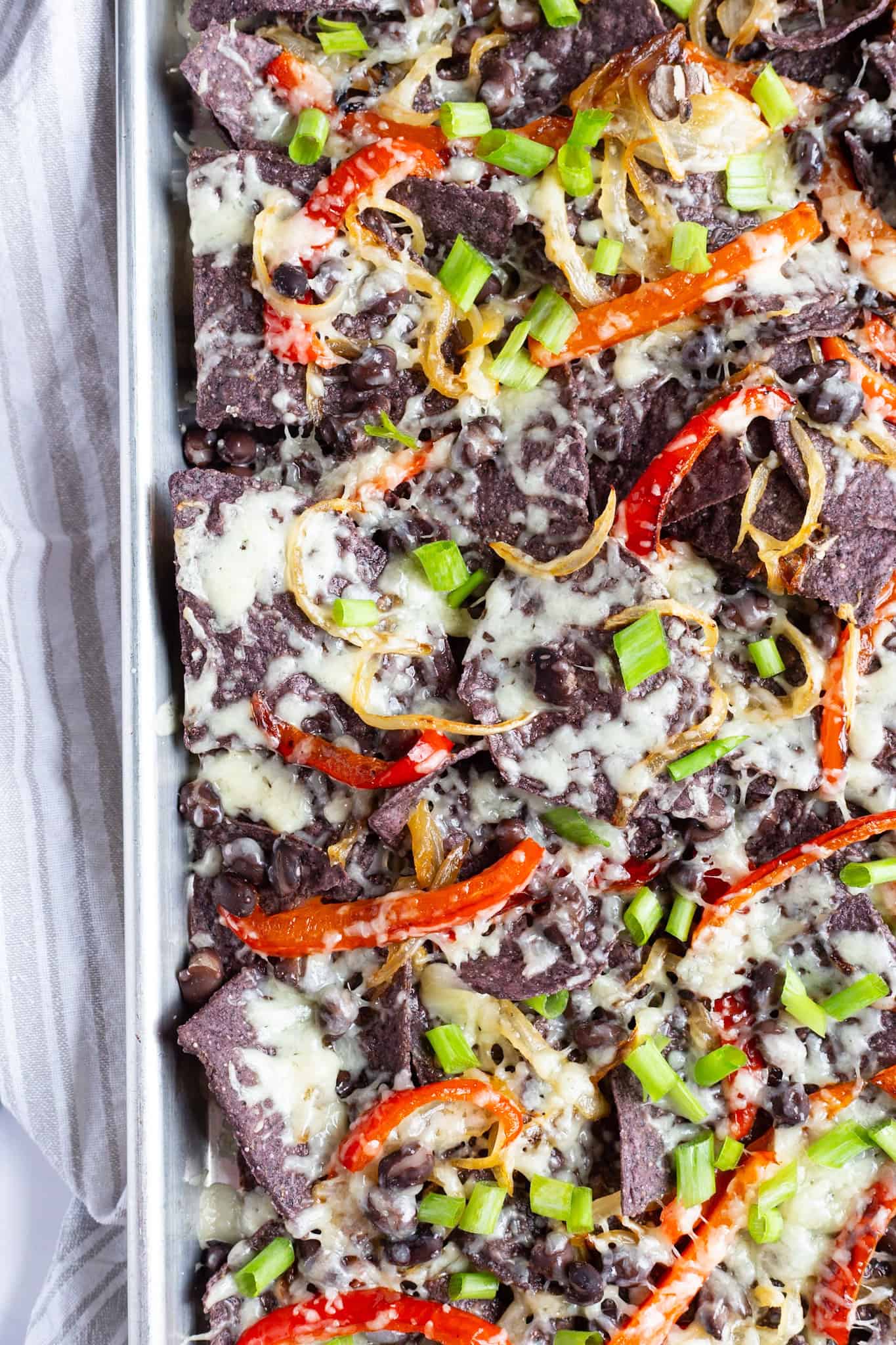 A colorful pan of veggie nachos featuring blue corn tortilla chips and topped with peppers, onions, black beans, scallions, and white cheddar.