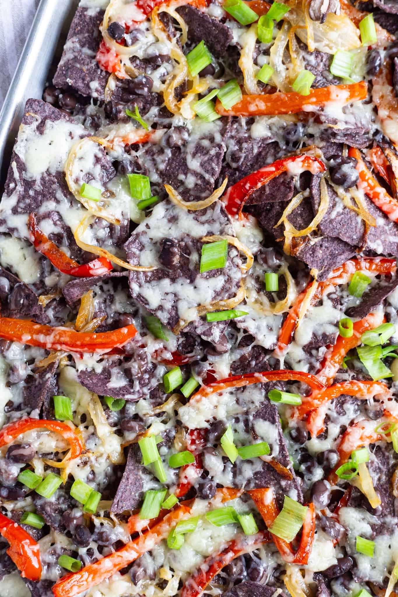 A sheet pan filled with blue tortilla chips and topped with red bell peppers, onions, black beans, scallions, and melted white cheddar.