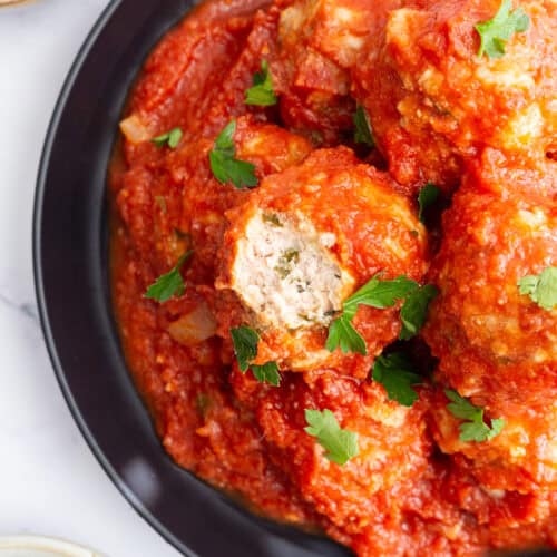 A dark plate filled with meatballs, with a bit taken from one to show the tender inside.