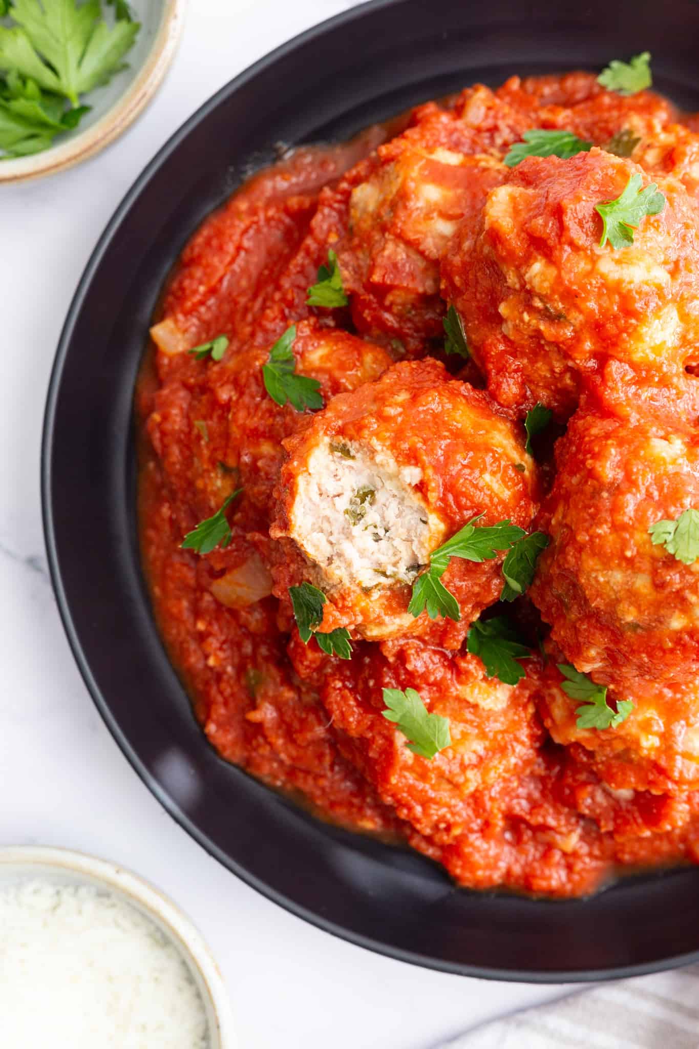 A dark plate filled with meatballs in marinara sauce, with a bite taken from one to show the tender inside.